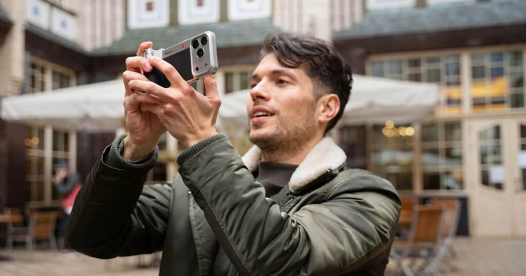 A man in an olive green jacket holds an iPhone equipped with a silver and black vintage-style camera grip. He is holding the device horizontally to take a photo in an outdoor courtyard with cafe seating and historic architecture in the background.