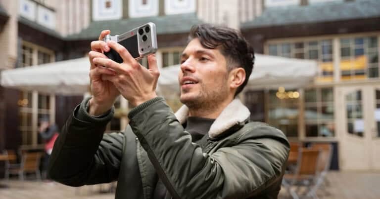 A man in an olive green jacket holds an iPhone equipped with a silver and black vintage-style camera grip. He is holding the device horizontally to take a photo in an outdoor courtyard with cafe seating and historic architecture in the background.