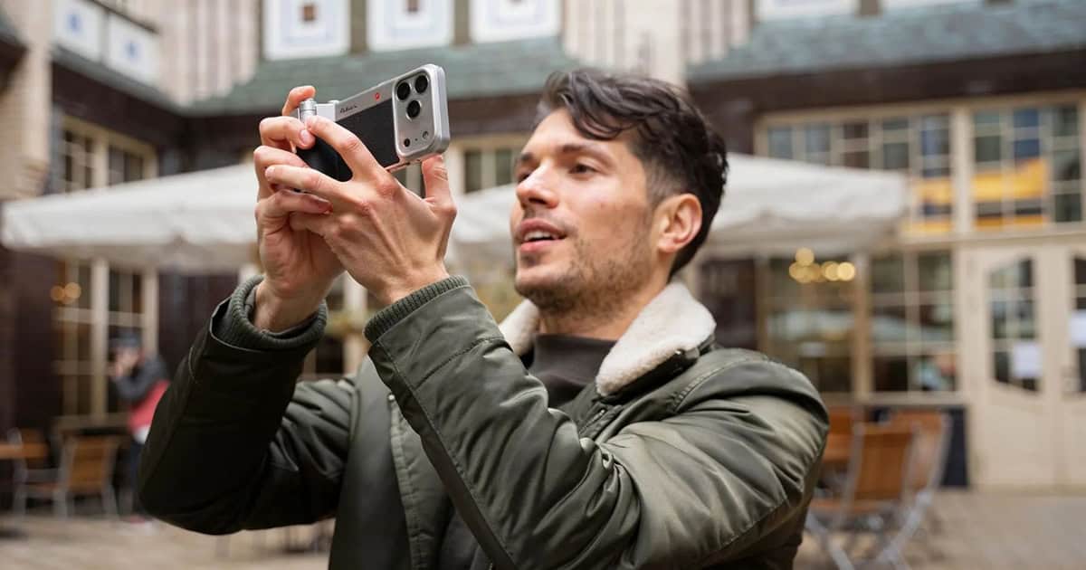 A man in an olive green jacket holds an iPhone equipped with a silver and black vintage-style camera grip. He is holding the device horizontally to take a photo in an outdoor courtyard with cafe seating and historic architecture in the background.
