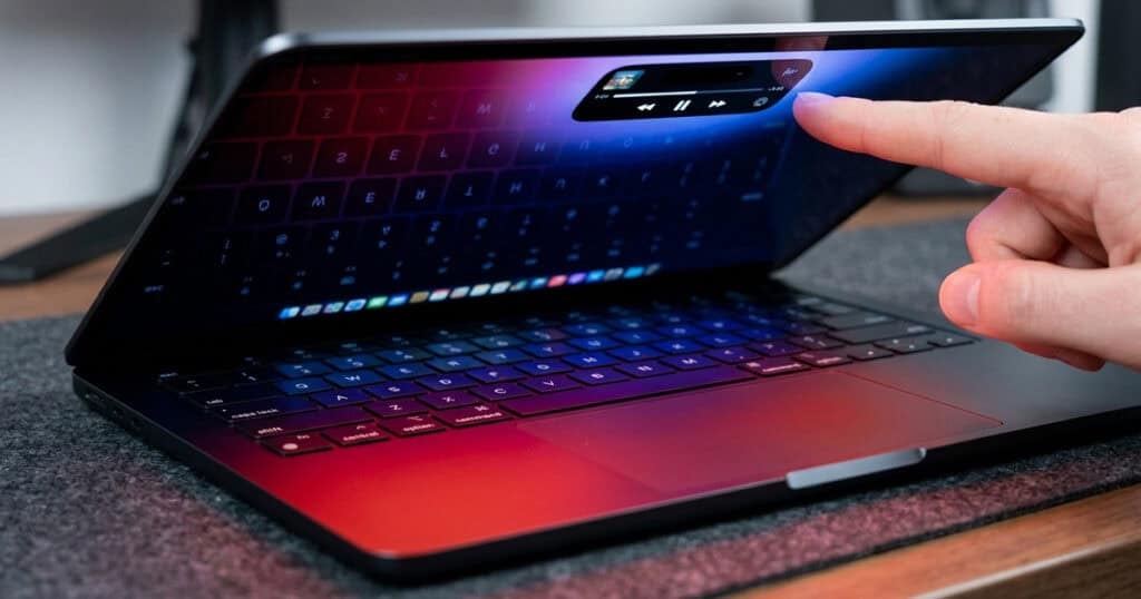 A finger reaching out to tap a floating media playback widget on the screen of a partially closed MacBook Pro, with a red and blue gradient wallpaper reflecting the keyboard below.
