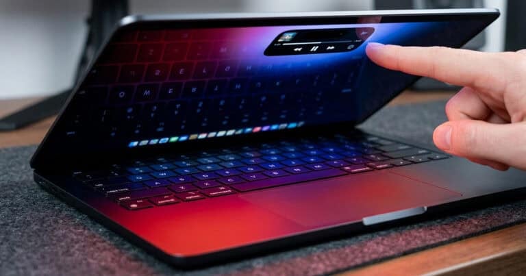 A finger reaching out to tap a floating media playback widget on the screen of a partially closed MacBook Pro, with a red and blue gradient wallpaper reflecting the keyboard below.